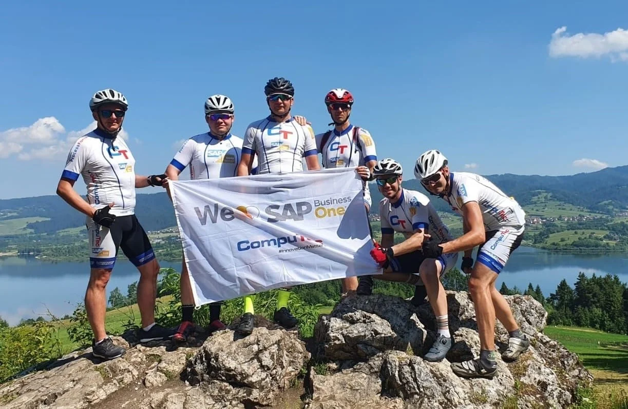 Six CompuTec team members holding a 'We love SAP Business One' flag on a mountain summit during a company cycling trip