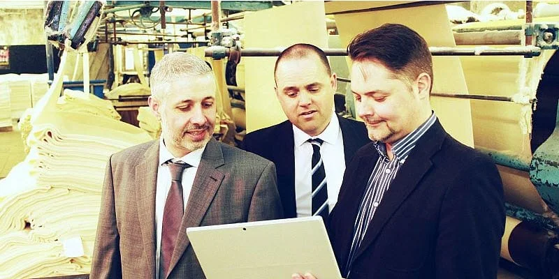 Three men in business suits reviewing a tablet on the William Clark fabric production floor